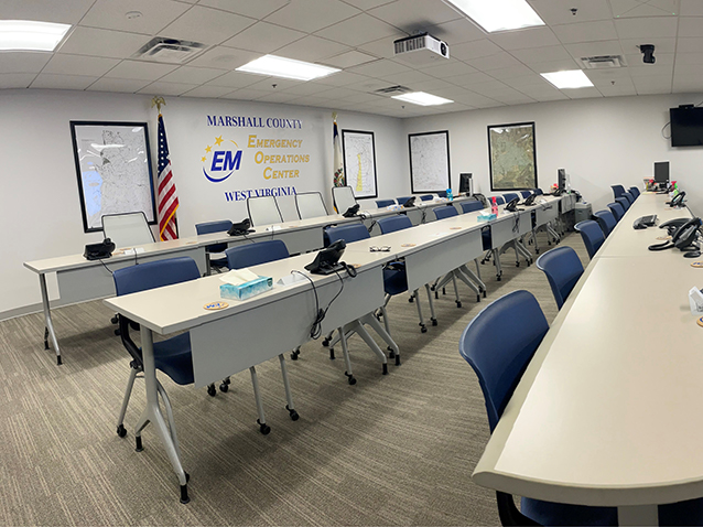 Interior of the Marshall County WV Emergency Operations Center with rows of empty tables and chairs, multiple monitors and informational posters on the walls, and a presentation screen displaying the center's name.