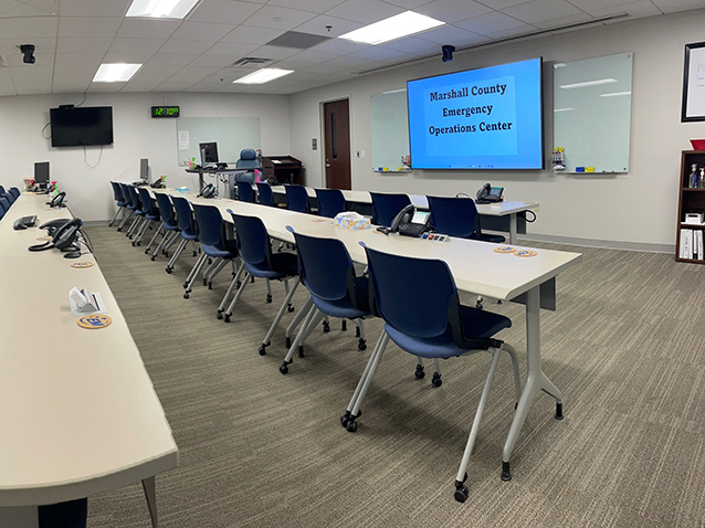 An interior view of the Marshall County Emergency Operations Center featuring a conference room setup with rows of empty chairs facing a projector screen displaying the center's name.